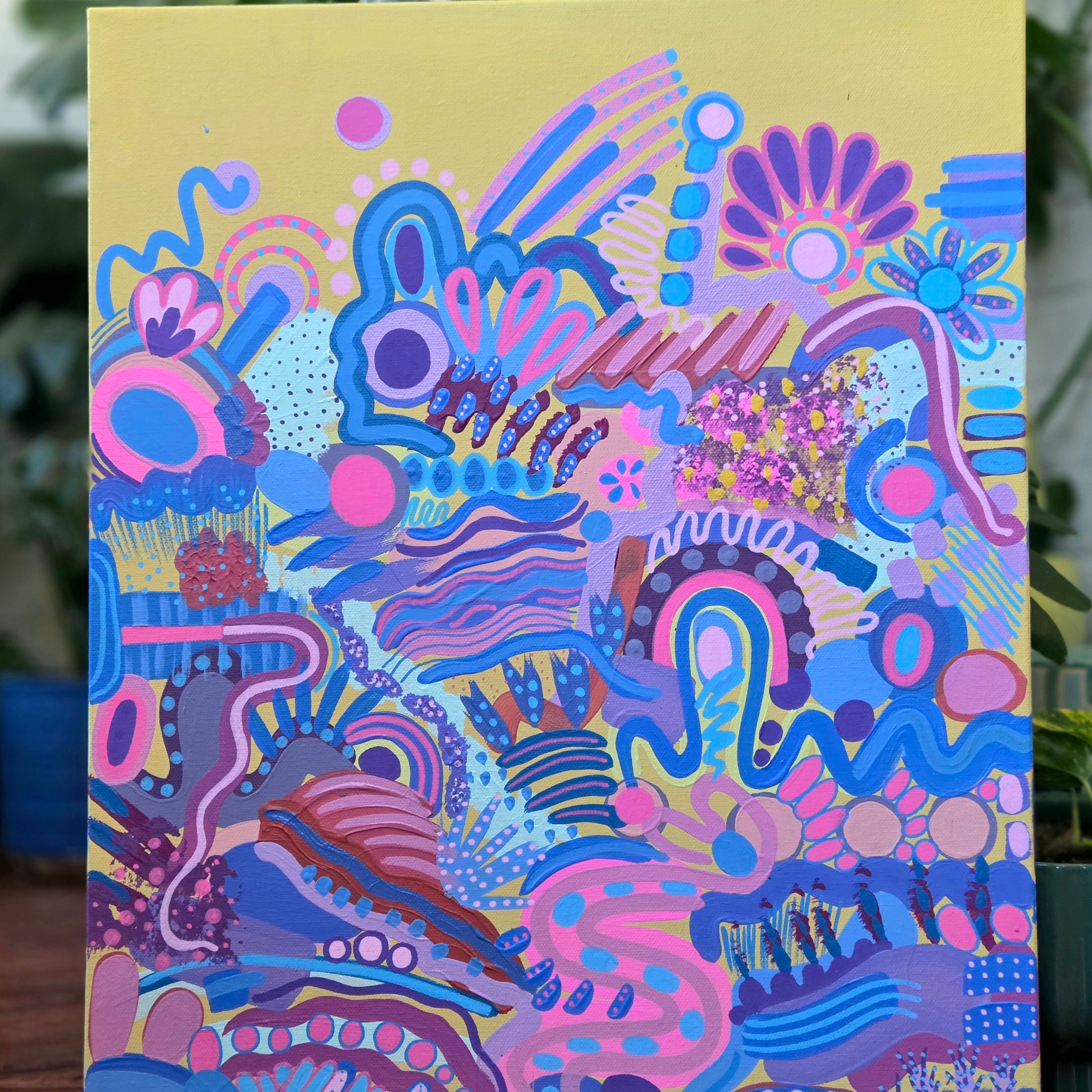 A colorful abstract canvas painting featuring vibrant pink, blue, purple, and yellow organic patterns, displayed on a wooden surface with green plants in the background.