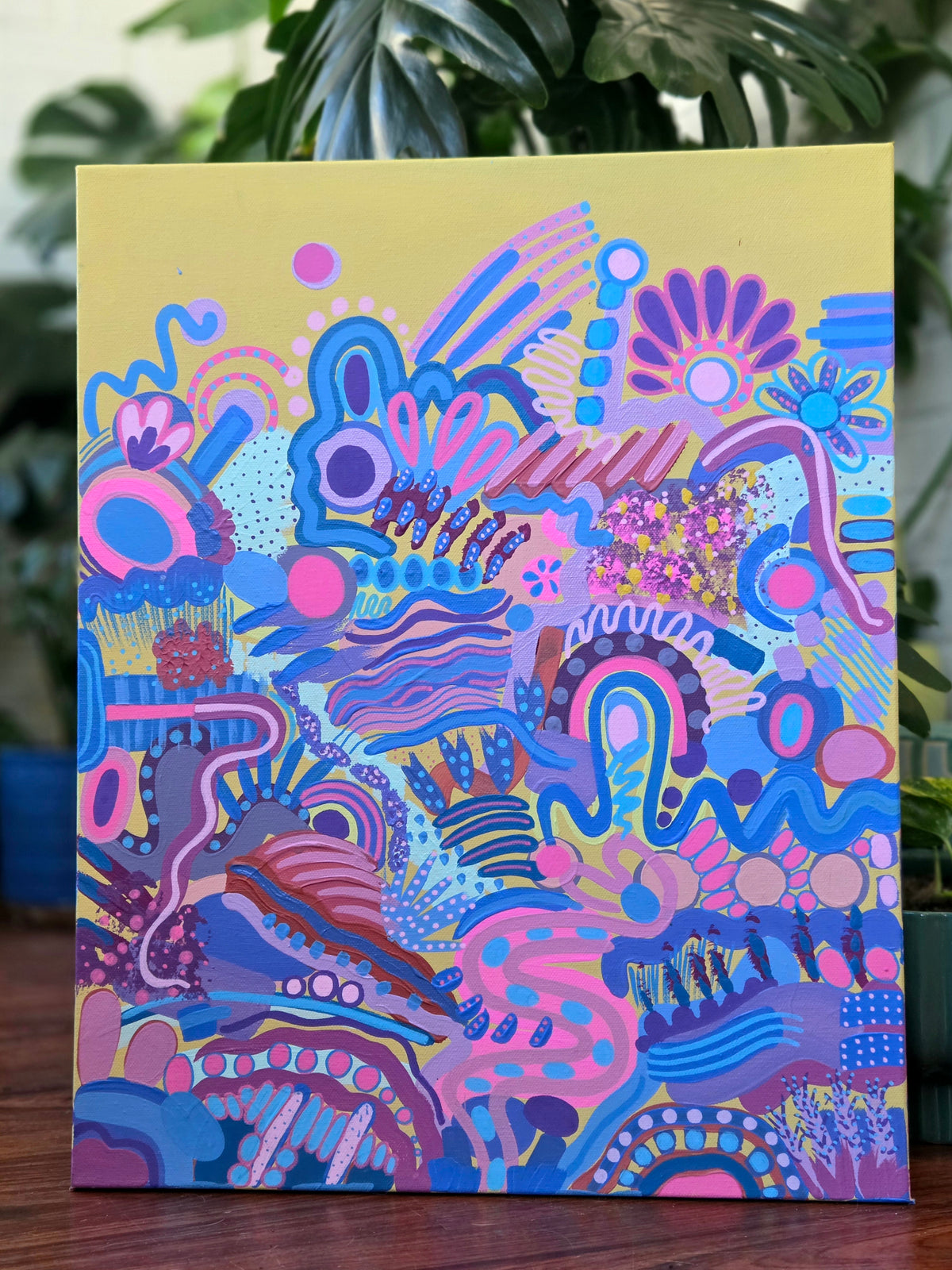 A colorful abstract canvas painting featuring vibrant pink, blue, purple, and yellow organic patterns, displayed on a wooden surface with green plants in the background.